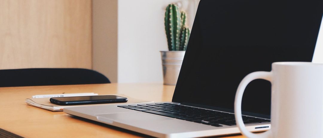 Ein offener Laptop auf einem Holztisch, ein Telefon, eine Tasse und ein Kaktus in einem Topf. Die Wand und ein Holzschrank sind im Hintergrund zu sehen.