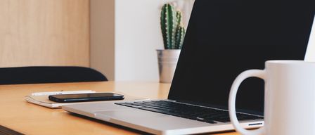 Ein offener Laptop auf einem Holztisch, ein Telefon, eine Tasse und ein Kaktus in einem Topf. Die Wand und ein Holzschrank sind im Hintergrund zu sehen.