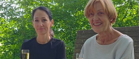 Zwei Frauen sitzen auf einer Bank im Park, lächeln und halten Weingläser. Die Frau auf der linken Seite trägt ein schwarzes Oberteil und Hose. Die Frau auf der rechten Seite trägt eine Uhr und ein weißes Hemd.