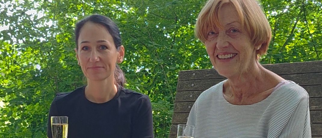 Zwei Frauen sitzen auf einer Bank im Park, lächeln und halten Weingläser. Die Frau auf der linken Seite trägt ein schwarzes Oberteil und Hose. Die Frau auf der rechten Seite trägt eine Uhr und ein weißes Hemd.