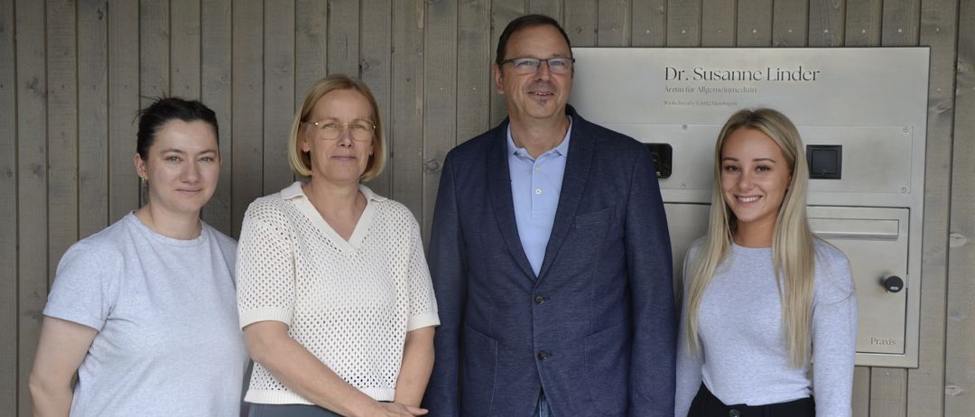 Vier Personen stehen zusammen vor einer Holzwand mit einem Schild, das Dr. Susanne Linde liest. Der Mann trägt einen blauen Anzug und die Frauen helle Blusen.