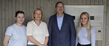 Vier Personen stehen zusammen vor einer Holzwand mit einem Schild, das Dr. Susanne Linde liest. Der Mann trägt einen blauen Anzug und die Frauen helle Blusen.