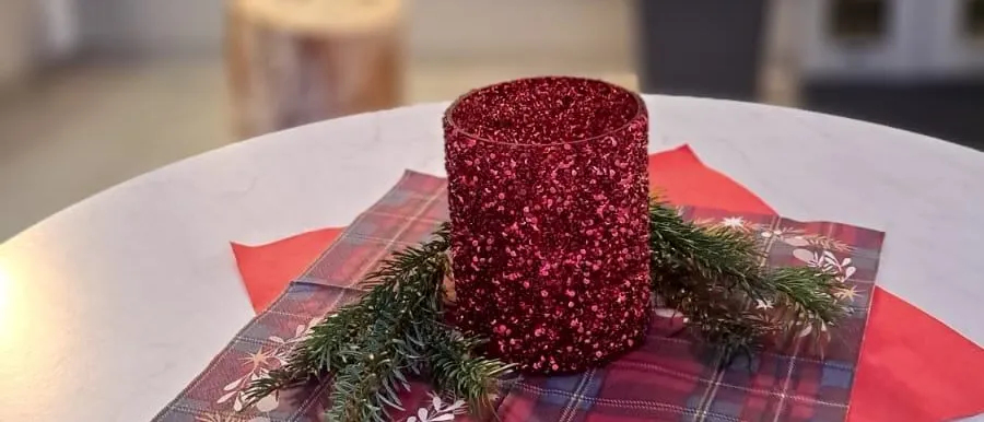 Eine rote glitzernde Kerze steht auf einem rot-schwarz karierten Tischset auf einem weißen Tisch, mit einem Topf und Glasfenstern im Hintergrund.
