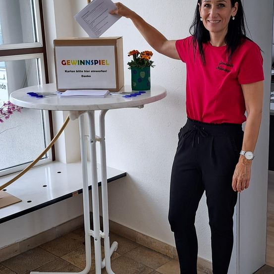 Eine Frau in einem roten T-Shirt und schwarzen Hosen steht neben einem Tisch mit einer Box und einer Pflanze. Sie hält ein Stück Papier und lächelt. Die Box hat bunten Text und Blumen darin.