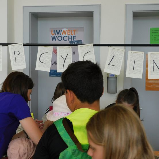 In einem Raum versammeln sich Kinder und Erwachsene um eine Wand mit einem Banner, das den Text 'Umwelt Woche' trägt. Die Wand hat zwei Türen, und mehrere Papiere mit Buchstaben hängen an einer Stange. Die Leute schauen sich die Papiere an.