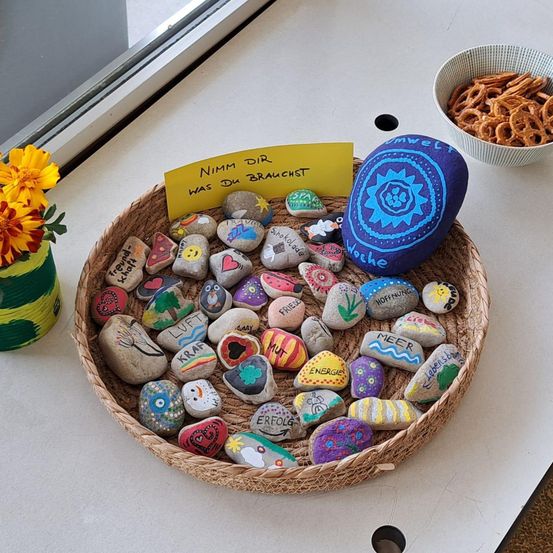 Ein Korb mit bemalten Steinen und einem gelben Schild mit der Aufschrift Nimm dir was du brauchst. Ein Schüssel mit Brezeln befindet sich rechts. Eine gelbe Blume in einem grünen Glas steht links.