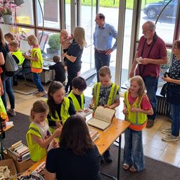 Eine Gruppe von Kindern und Erwachsenen in gelben Westen steht um einen Tisch mit Büchern. Eine Frau hält ein Baby, während ein Mann ein Papier liest. Sie befinden sich in einem Flur mit Glastüren.