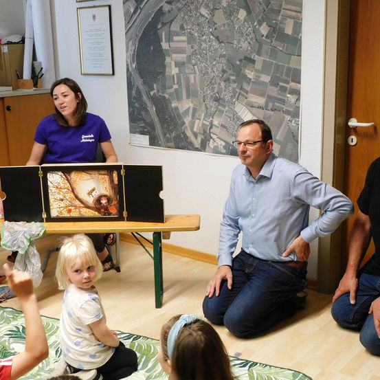 Eine Gruppe von Menschen, darunter eine Frau und ein Mann, sitzen auf dem Boden um einen Tisch mit einem Bilderbuch. Sie sind drinnen in einem Raum mit Holzboden und einem großen Luftbild an der Wand.