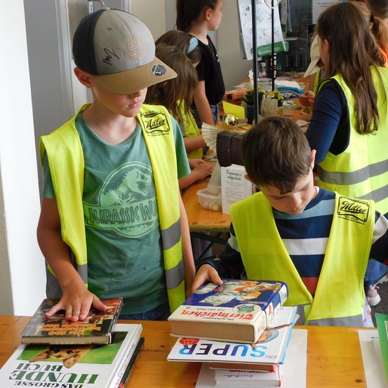 Zwei Jungen in Warnwesten konzentrieren sich auf Bücher an einem Tisch mit anderen dahinter in einem Raum.