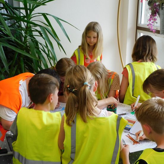 Eine Gruppe von Kindern in Sicherheitswesten sitzt an einem Tisch und zeichnet und malt mit Markern. Ein Mädchen steht hinter ihnen und schaut sich etwas an.