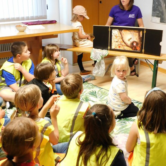 Kinder in gelben Warnwesten versammeln sich um eine Frau, die ein Bild auf einem Tisch zeigt, ein Kind schaut in die Kamera.