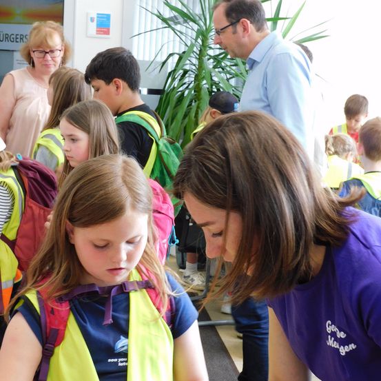 Eine Gruppe von Kindern mit Sicherheitswesten und Rucksäcken versammelt sich in einem Gebäude. Eine Frau in einem lila Shirt beugt sich vor, um einem Mädchen in einer gelben Weste zu helfen. Im Hintergrund beobachten Erwachsene die Szene.