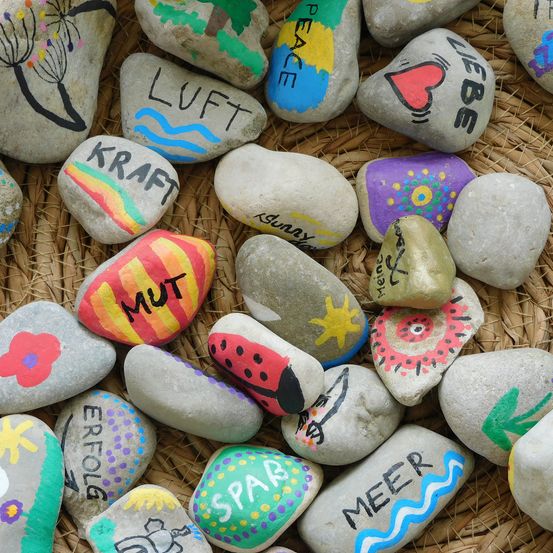 Ein Korb aus Weide geflochten, gefüllt mit Steinen verschiedener Größen und Farben. Jeder Stein ist mit verschiedenen Symbolen, Wörtern und Zeichnungen bemalt. Wörter beinhalten 'frieden', 'liebe', 'kraft', 'mut' und 'meer'. Symbole umfassen ein Herz, einen Baum, einen Löwenzahn und eine Marienkäfer.