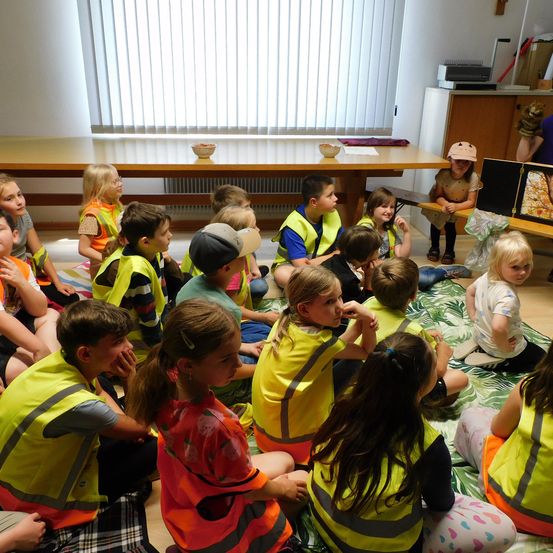 Eine Gruppe von Kindern in Sicherheitswesten sitzt im Kreis auf dem Boden, während eine Frau in der Nähe ein Buch liest.