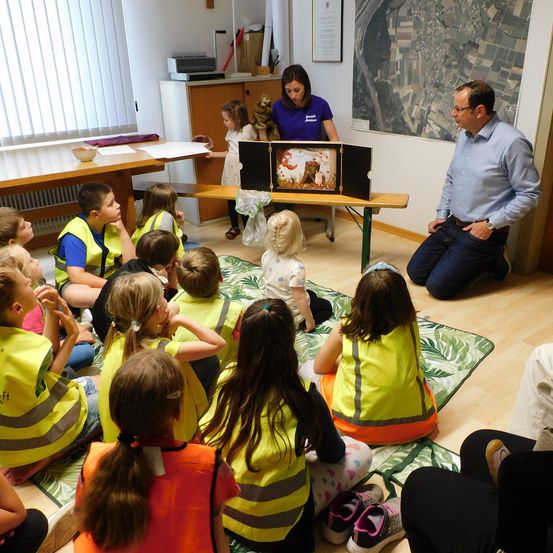 Eine Gruppe von Kindern in Sicherheitswesten sitzt auf einem Teppich, während eine Frau in einem blauen Shirt ein offenes Buch mit einem Bild hält. Ein Mann in einem blauen Shirt kniet neben ihnen.