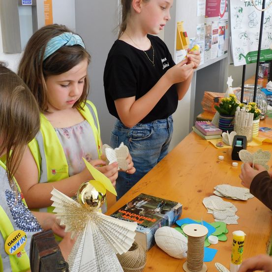Drei Mädchen sind bei einer Aktivität an einem Tisch beschäftigt. Ein Mädchen trägt eine gelbe Weste und schaut sich einen Papierengel an. Ein anderes Mädchen trägt ein schwarzes Shirt und hält ein gelbes Objekt. Verschiedene Bastelmaterialien liegen auf dem Tisch.