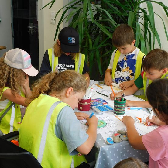 Eine Gruppe von Kindern in Sicherheitswesten sitzt um einen Tisch herum und ist mit Malaktivitäten beschäftigt. Sie benutzen farbige Stifte und Marker. Eine große Pflanze steht hinter ihnen.