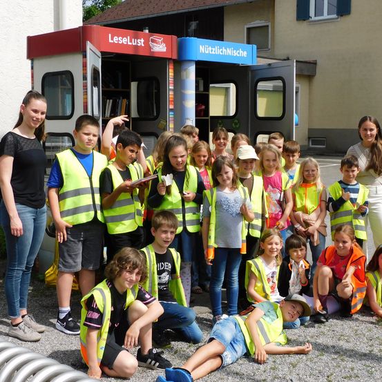 Eine Gruppe von Kindern in Warnwesten steht vor einem Bücherwagen und einem Haus, lächelt und posiert für ein Foto.
