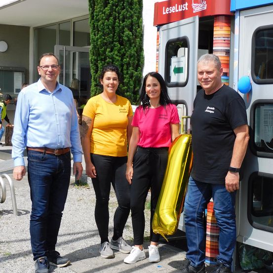 Vier Menschen stehen vor einem bunten Gebäude mit einem Schild, auf dem LeseLust steht. Der Mann auf der linken Seite trägt ein hellblaues Hemd und eine Brille, während der Mann auf der rechten Seite ein schwarzes Hemd und eine Uhr trägt. Die beiden Frauen tragen gelbe und rosa Hemden und eine von ihnen hält einen goldenen Luftballon.