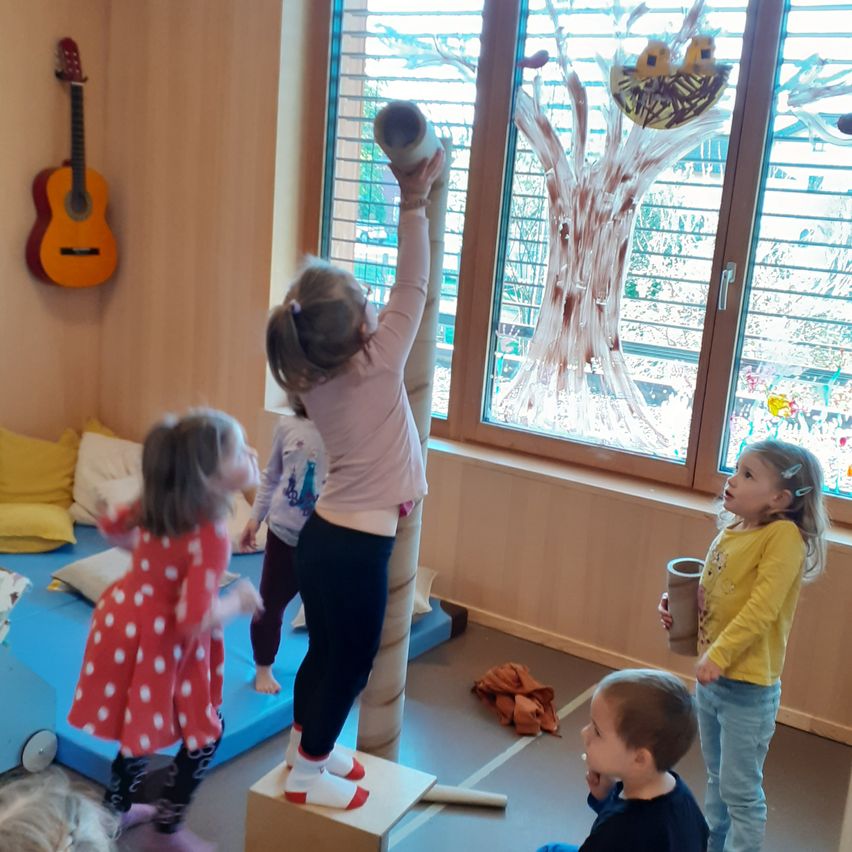 In einem Spielzimmer klettert ein kleines Mädchen auf eine Pappröhre, während andere zusehen. Ein Baumbild schmückt das Fenster, und eine Gitarre hängt an der Wand.