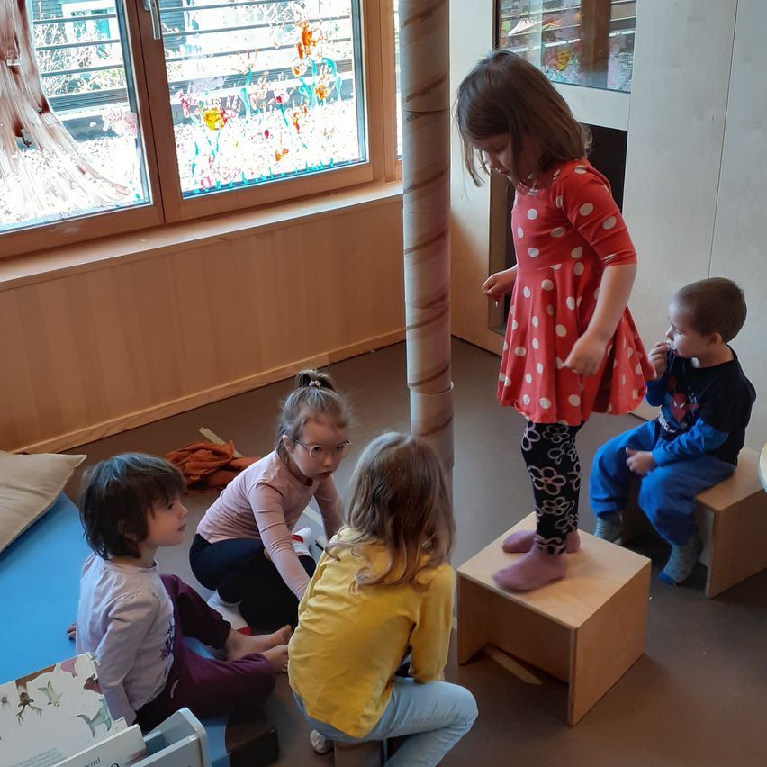 Eine Gruppe von Kindern spielt in einem Raum. Ein Kind steht auf einem Hocker, während andere um sie herumsitzen. Bunte Fenster und Holzmöbel sind im Hintergrund sichtbar.