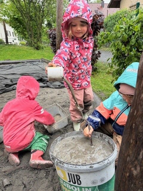Drei kleine Kinder in Regenmänteln mischen Zement in einem Eimer auf einem Spielplatz. Ein Kind hält einen Becher.