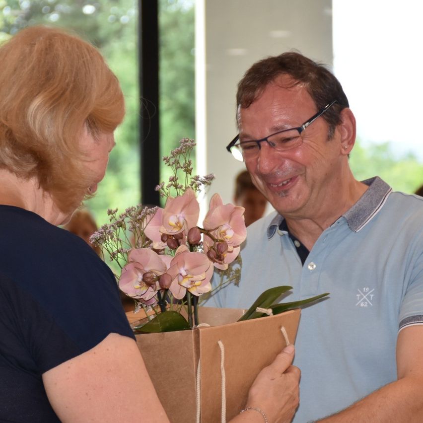 Ein lächelnder Mann in einem blauen Polohemd überreicht einer Frau mit blonden Haaren Blumen. Die Blumen sind pinkfarbene Orchideen in einem braunen Papierbeutel.