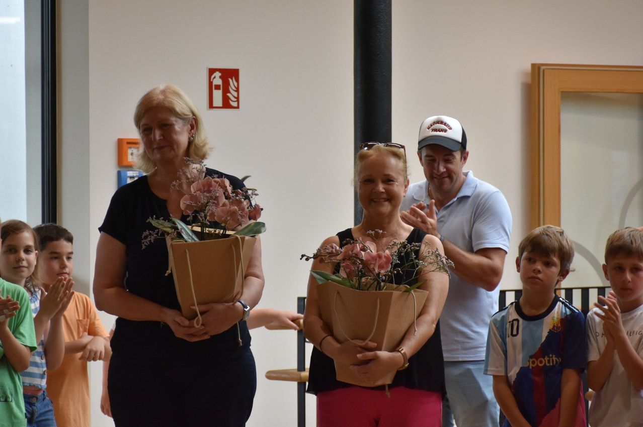 Zwei Frauen und ein Mann, die Blumensträuße halten, stehen zusammen, eine Frau lächelt, der Mann trägt eine Kappe, in einem Raum mit einem Feuerlöscher-Schild an der Wand.