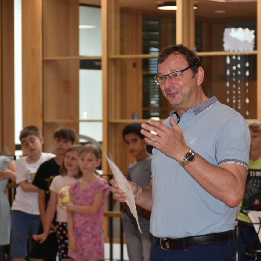 Ein Mann mit Brille hält eine Rede vor einer Gruppe von Kindern. Er hält ein Stück Papier in der Hand und macht Handbewegungen. Die Kinder stehen hinter ihm und schauen zu. Der Ort ist drinnen mit Holzregalen im Hintergrund.