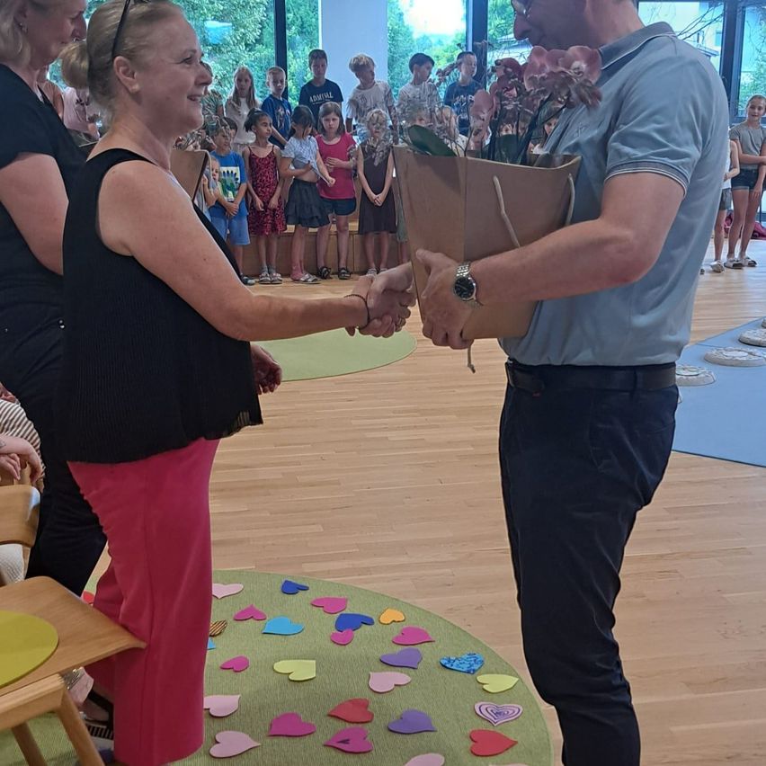 Eine Frau in rosa Hosen schüttelt einem Mann die Hand, der Blumen hält, während Kinder auf einem Holzboden mit einem herzförmigen Teppich hinter ihnen stehen.