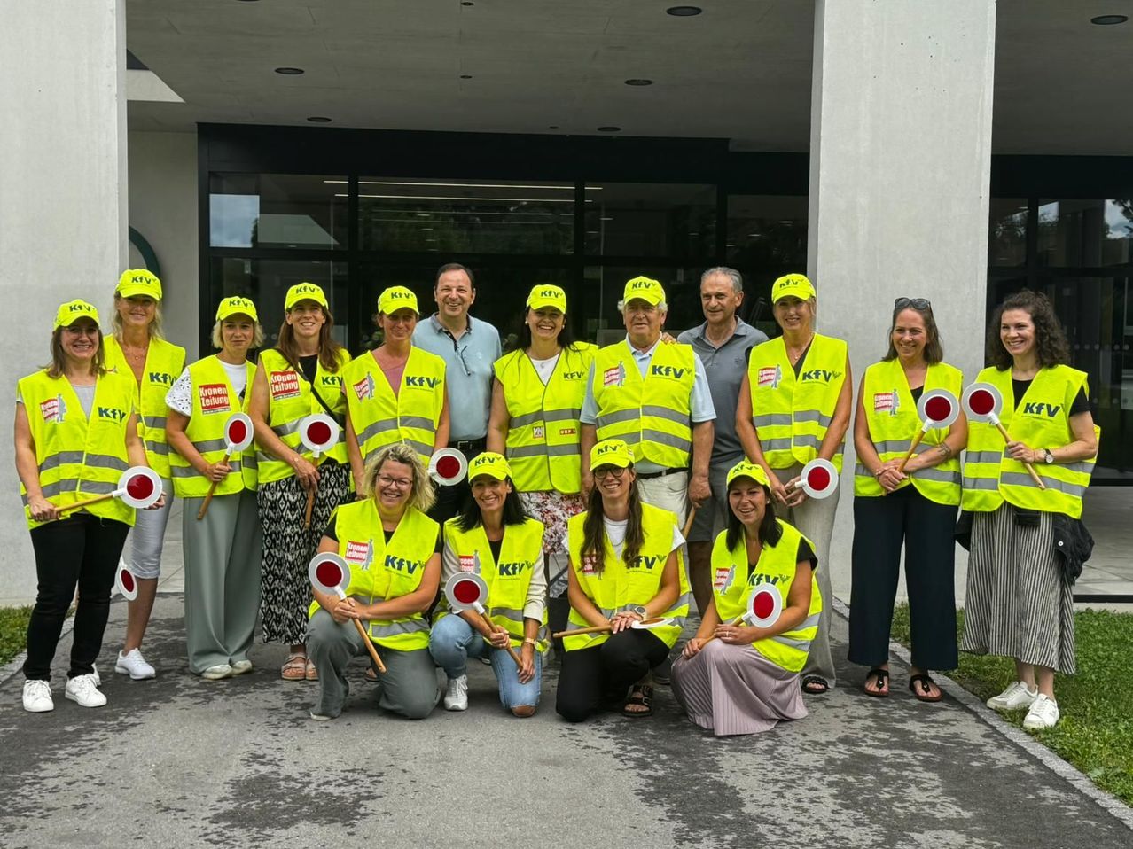 Eine Gruppe von Menschen in gelben Westen und Hüten posiert für ein Foto vor einem Gebäude. Sie halten rote Schilder mit weißen Kreisen.