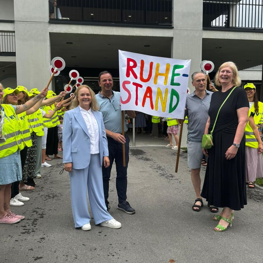 Eine Gruppe von Menschen steht vor einem Gebäude mit einem Schild, auf dem 'RUHE-STAND' steht. Sie tragen alle Warnwesten und halten Schilder.