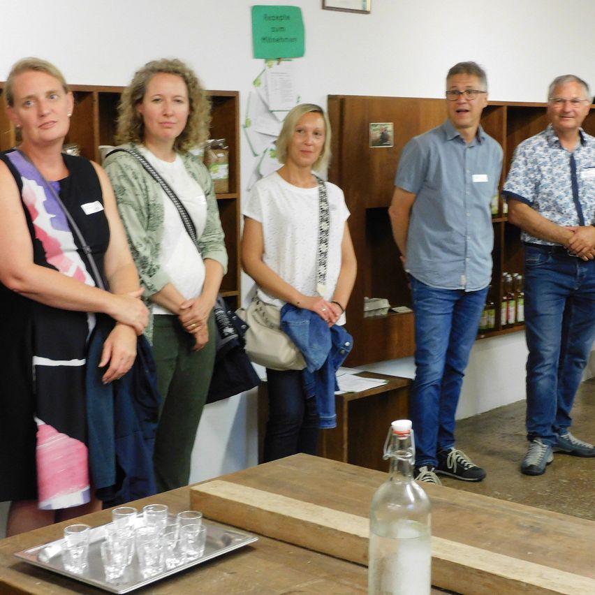Fünf Menschen stehen in einem Raum. Zwei Frauen und drei Männer. Sie haben die Hände in den Taschen. Vor ihnen steht eine Flasche und ein Tablett mit Gläsern.