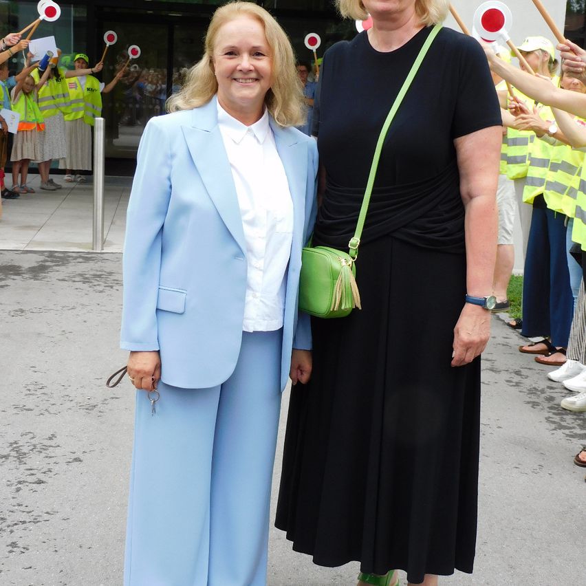 Zwei Frauen in Anzügen stehen lächelnd vor einem Gebäude. Sie sind von Menschen in gelben Westen umgeben, einige halten rote Flaggen.