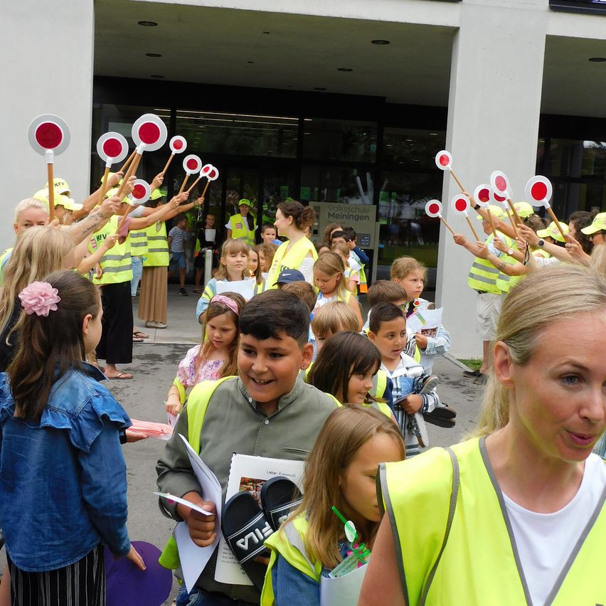 Eine Gruppe von Kindern und Erwachsenen in gelben Westen steht draußen und hält rote und weiße Schilder in der Hand. Einige der Kinder haben Hüte und Armbänder.