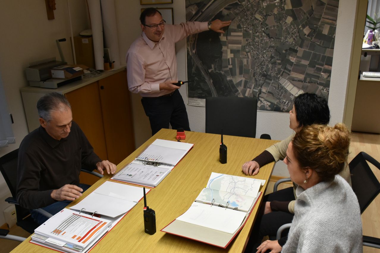 Ein Mann in einem rosa Hemd steht und zeigt auf eine große Karte an der Wand, wahrscheinlich in einer Besprechung. Mehrere Personen sitzen um einen Tisch mit Karten, Ordnern und Walkie-Talkies.