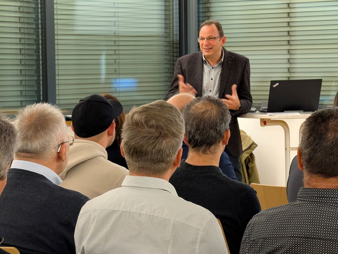 Ein Mann steht vor einer Gruppe von Menschen, spricht und gestikuliert mit den Händen. Das Publikum sieht ihn an. Hinter dem Mann steht ein Tisch mit einem Laptop darauf. Die Leute sitzen auf Stühlen. Der Raum hat Glasfenster mit Jalousien.