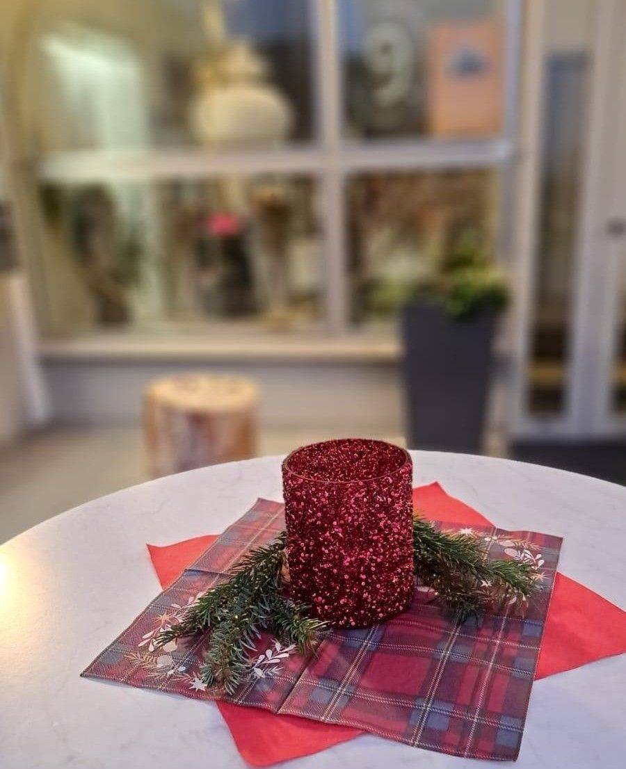 Eine rote glitzernde Kerze auf einem Tisch mit einem karierten Tuch und grünen Blättern, mit einem Holzhocker und einer Pflanze im Hintergrund.