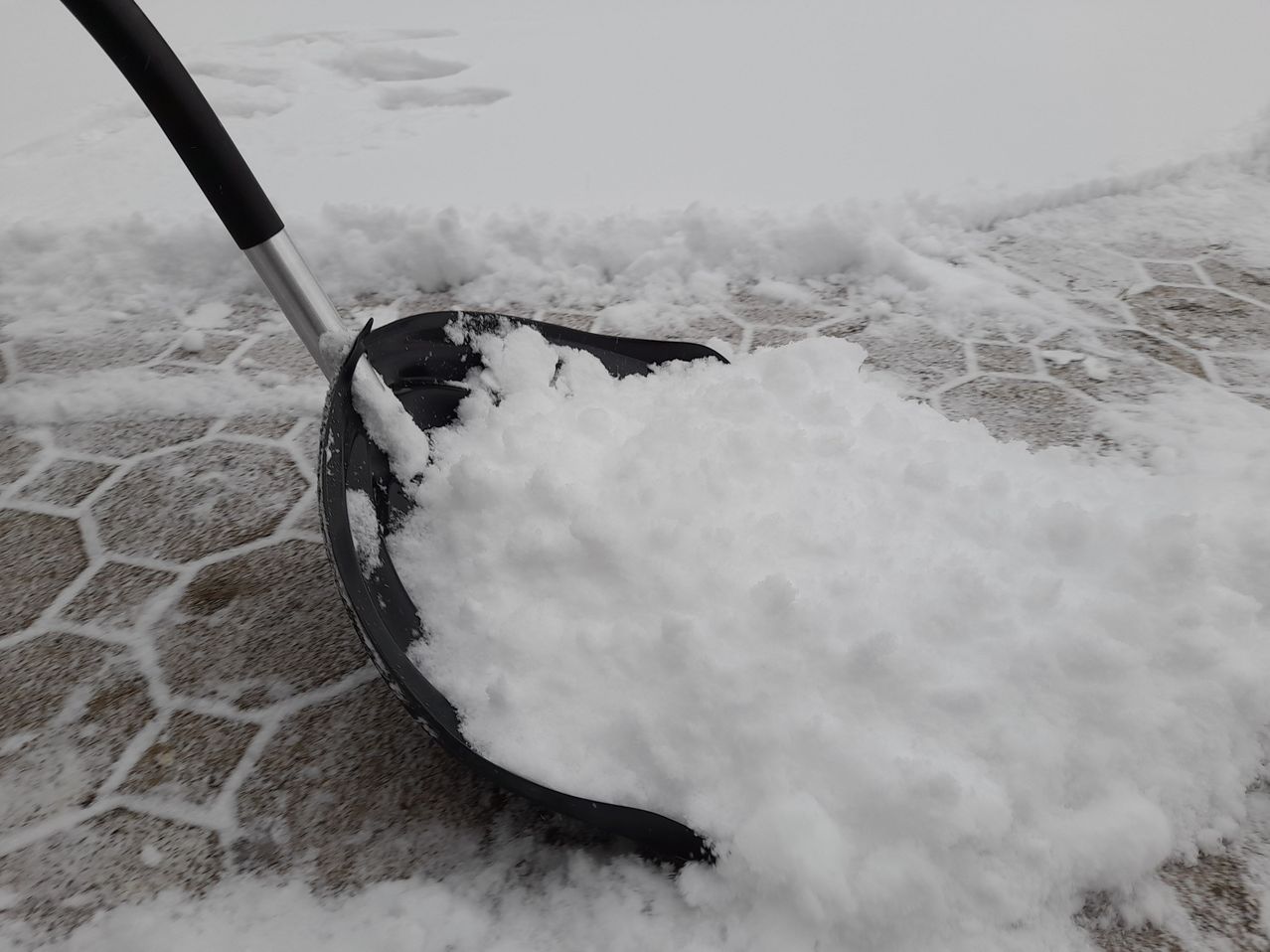 Eine schwarze Schneeschaufel voller Schnee steht auf einem grauen Fliesenboden, mit Spuren im Schnee dahinter.