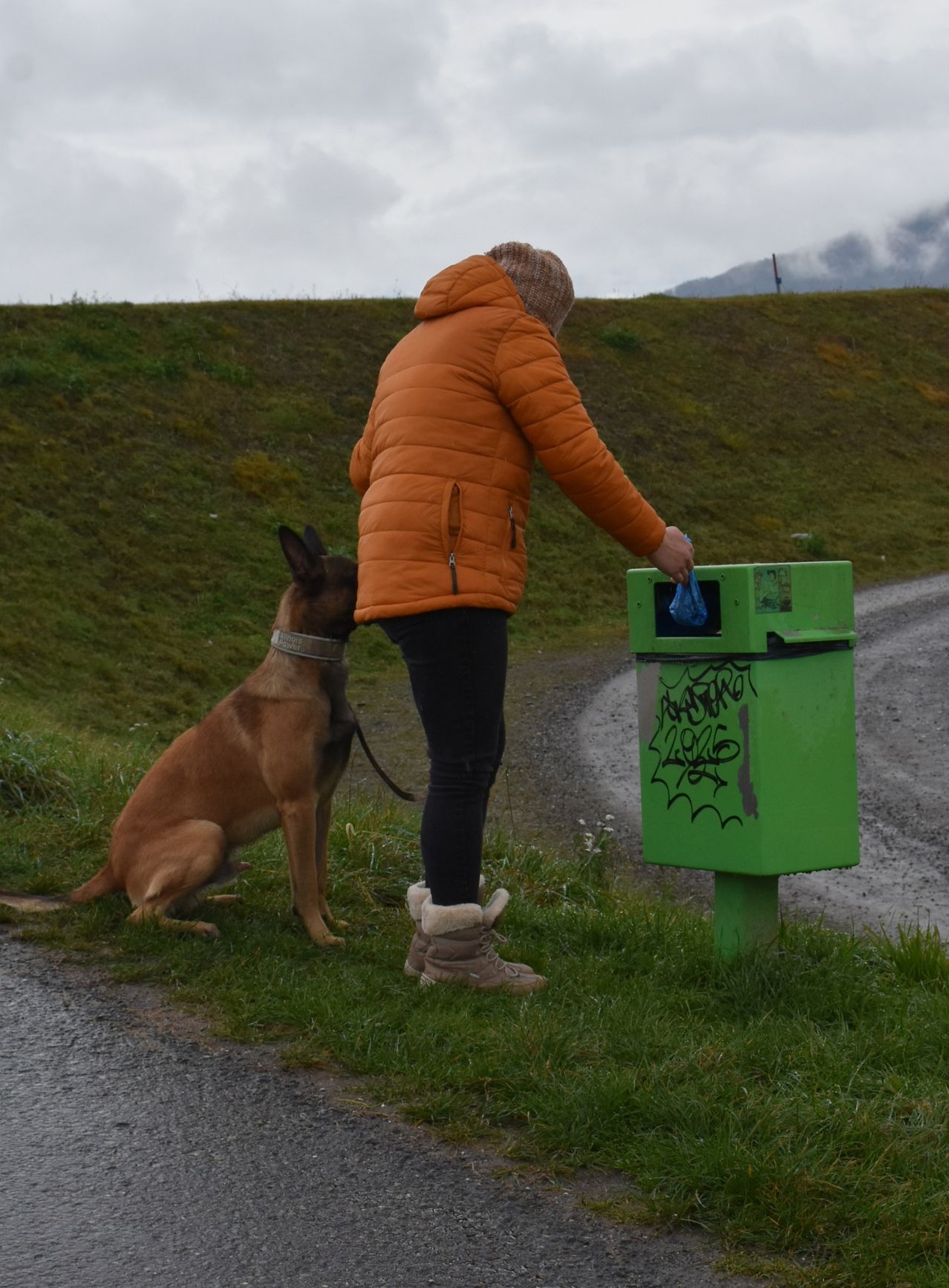 Eine Person in einer orangefarbenen Jacke entsorgt einen Plastikbeutel in einem grünen Abfallbehälter, während ihr Hund wartet.