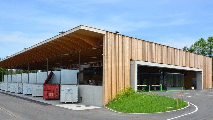 Eine moderne Einrichtung mit hölzerne Außenseite und großer offener Fläche, mehrere weiße Behälter und ein roter vorn, unter einem Dach mit Metallbalken, an einem sonnigen Tag.