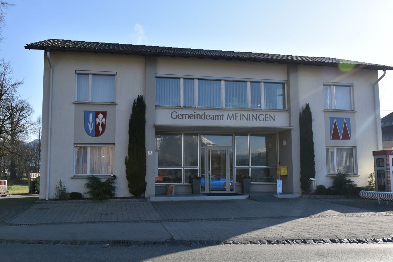 Das Gemeindeamt Meiningen befindet sich vor einem blauen Auto, das vor dem Gebäude mit Fenstern und Glastüren geparkt ist.