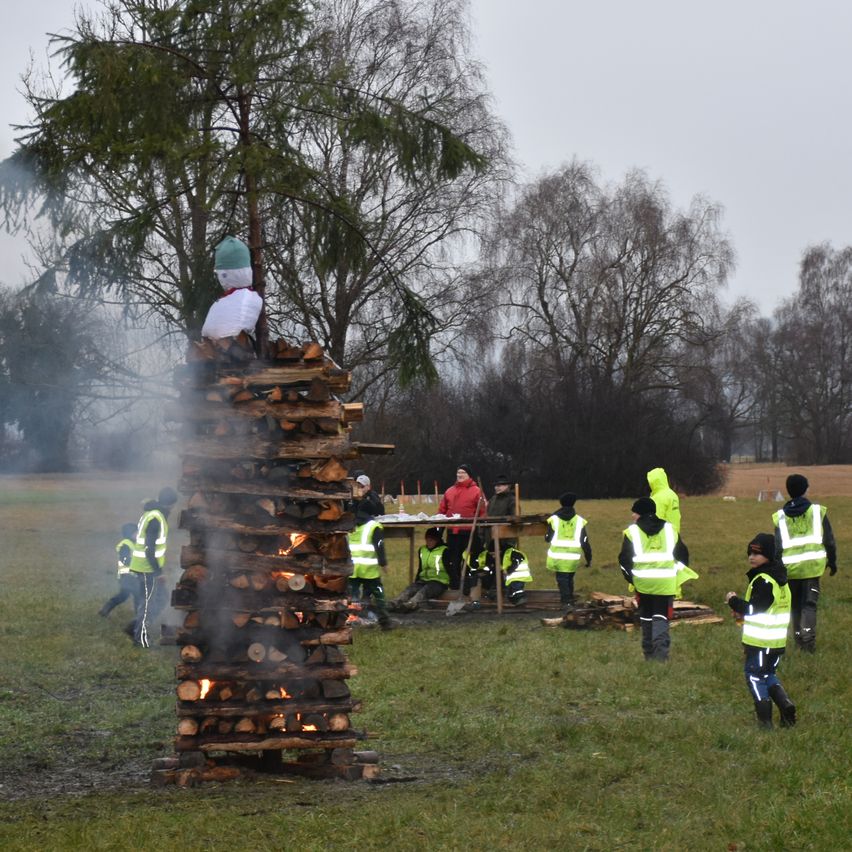 Eine Gruppe von Menschen versammelt sich um einen brennenden Holzstapel auf einer Wiese. Einige tragen Sicherheitswesten, während andere bei einem Tisch stehen. Bäume sind im Hintergrund.