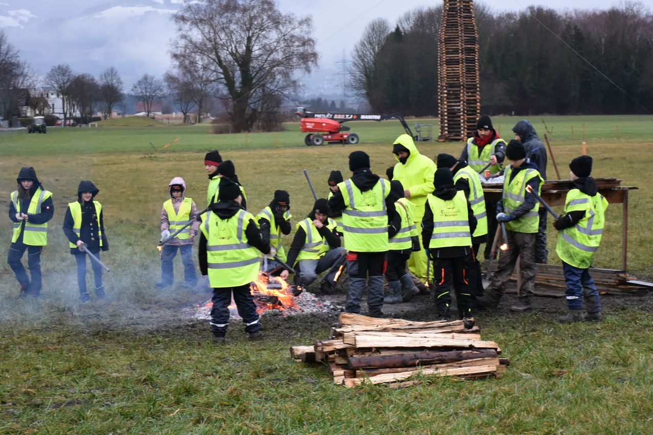 Eine Gruppe von Menschen in Sicherheitswesten steht um ein Lagerfeuer herum und bereitet sich auf ein Lagerfeuer-Event in einem Grasfeld vor. Im Hintergrund steht eine hohe Holzkonstruktion.
