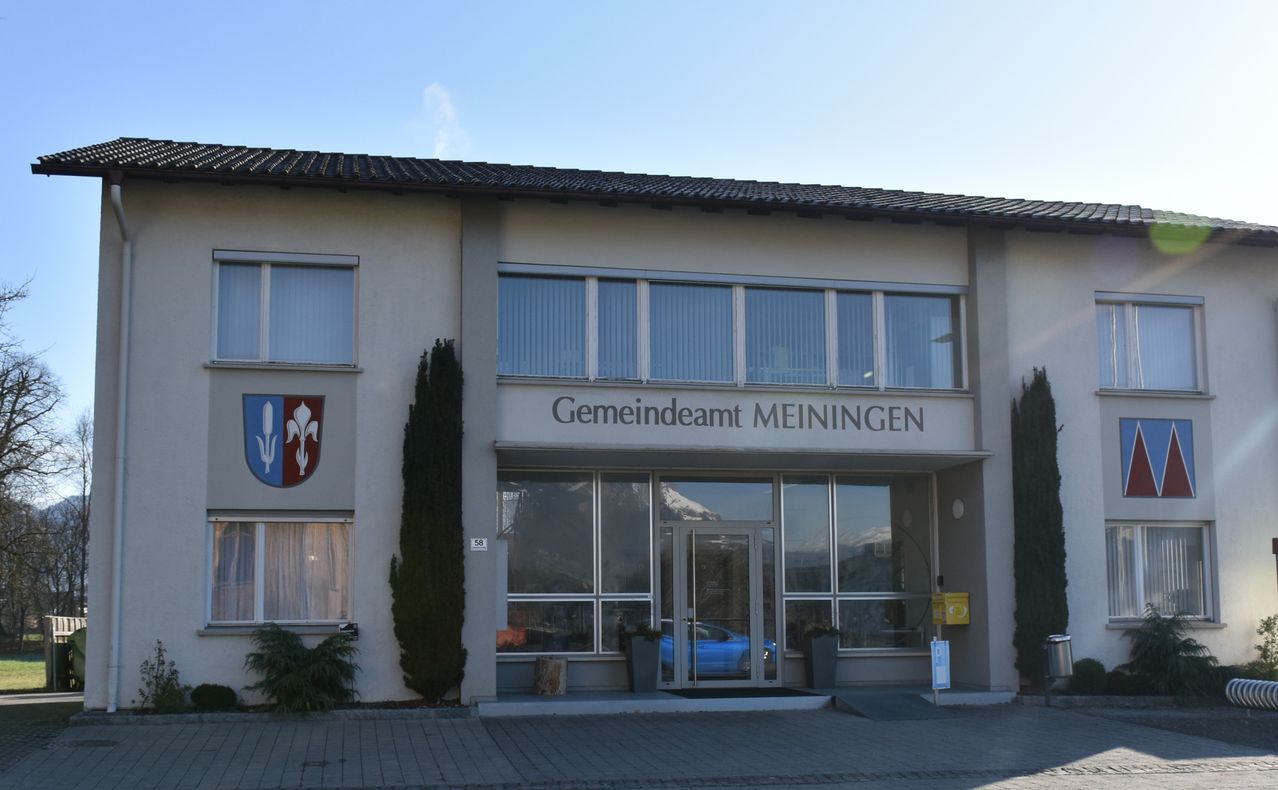 Die Vorderseite des Gemeindeamt Meiningen Gebäudes mit Glasfenstern, einem geparkten blauen Auto davor und einem Berg im Hintergrund.