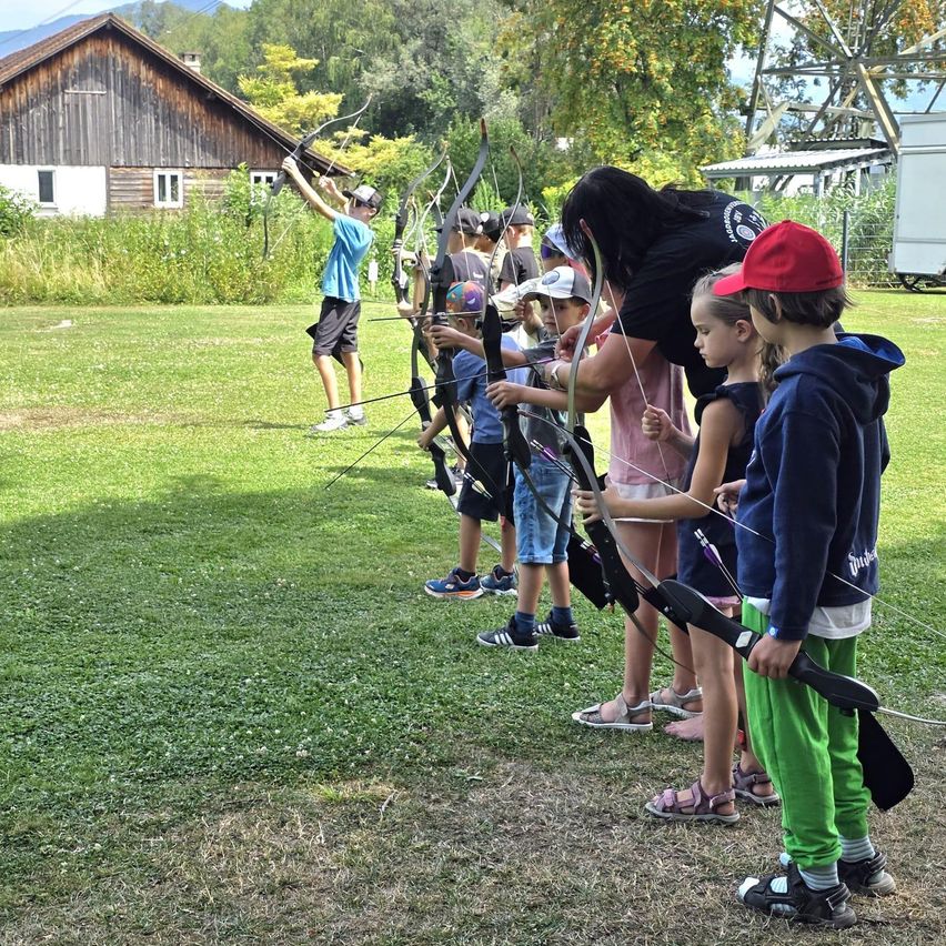 A group of children and adults are practicing archery on a grassy field, with a barn and trees in the background.