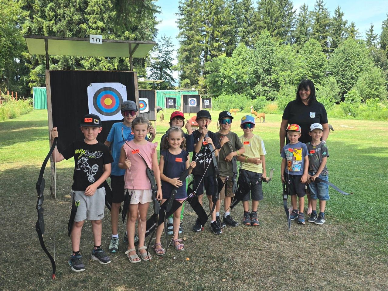 A group of children and an adult woman are standing in a field, holding bows and arrows, with a target board behind them.