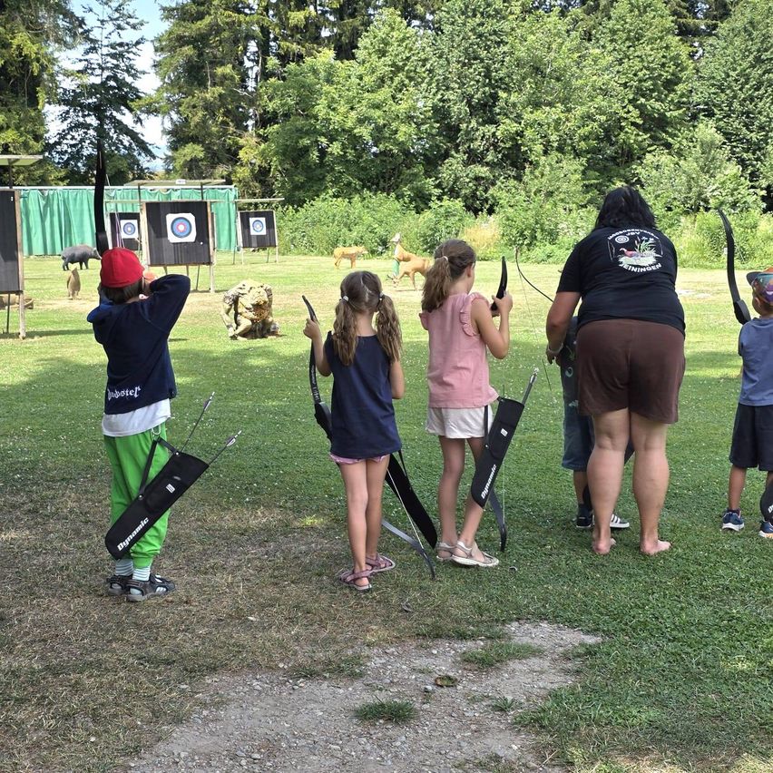 A group of children and adults are practicing archery in an outdoor setting with green trees and a grassy field.