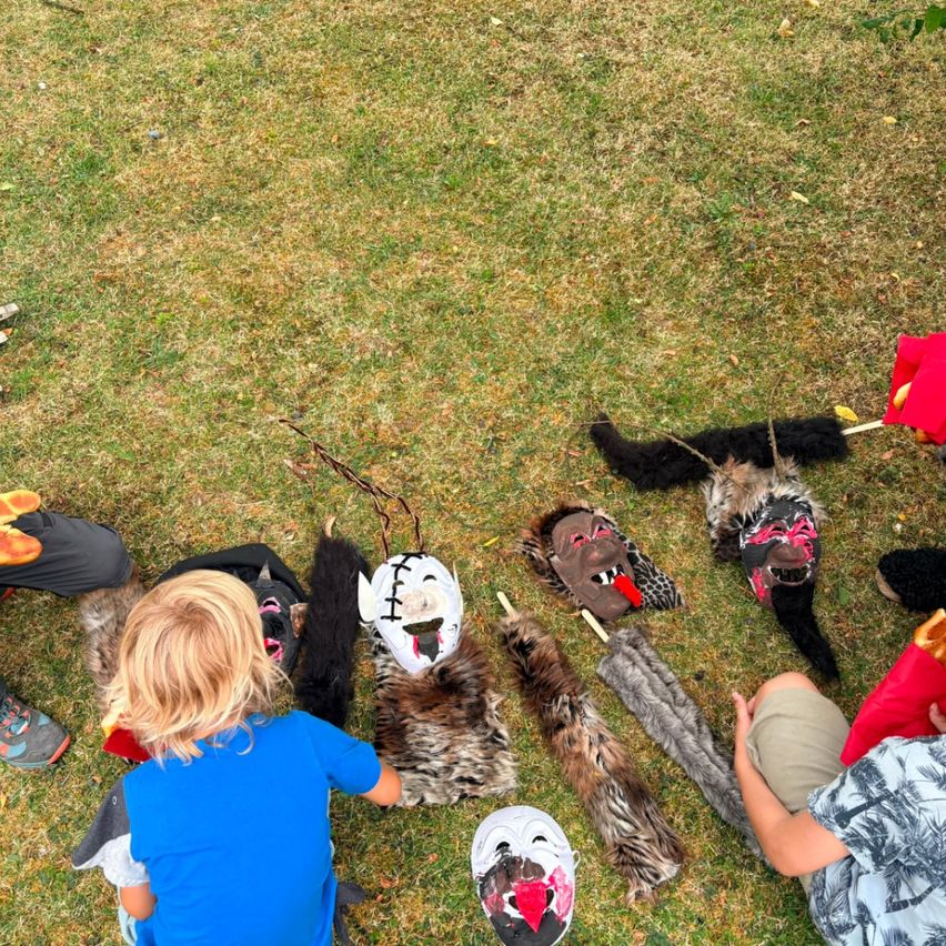 Children sit on grass with animal masks and tails around them, possibly preparing for a play or performance.