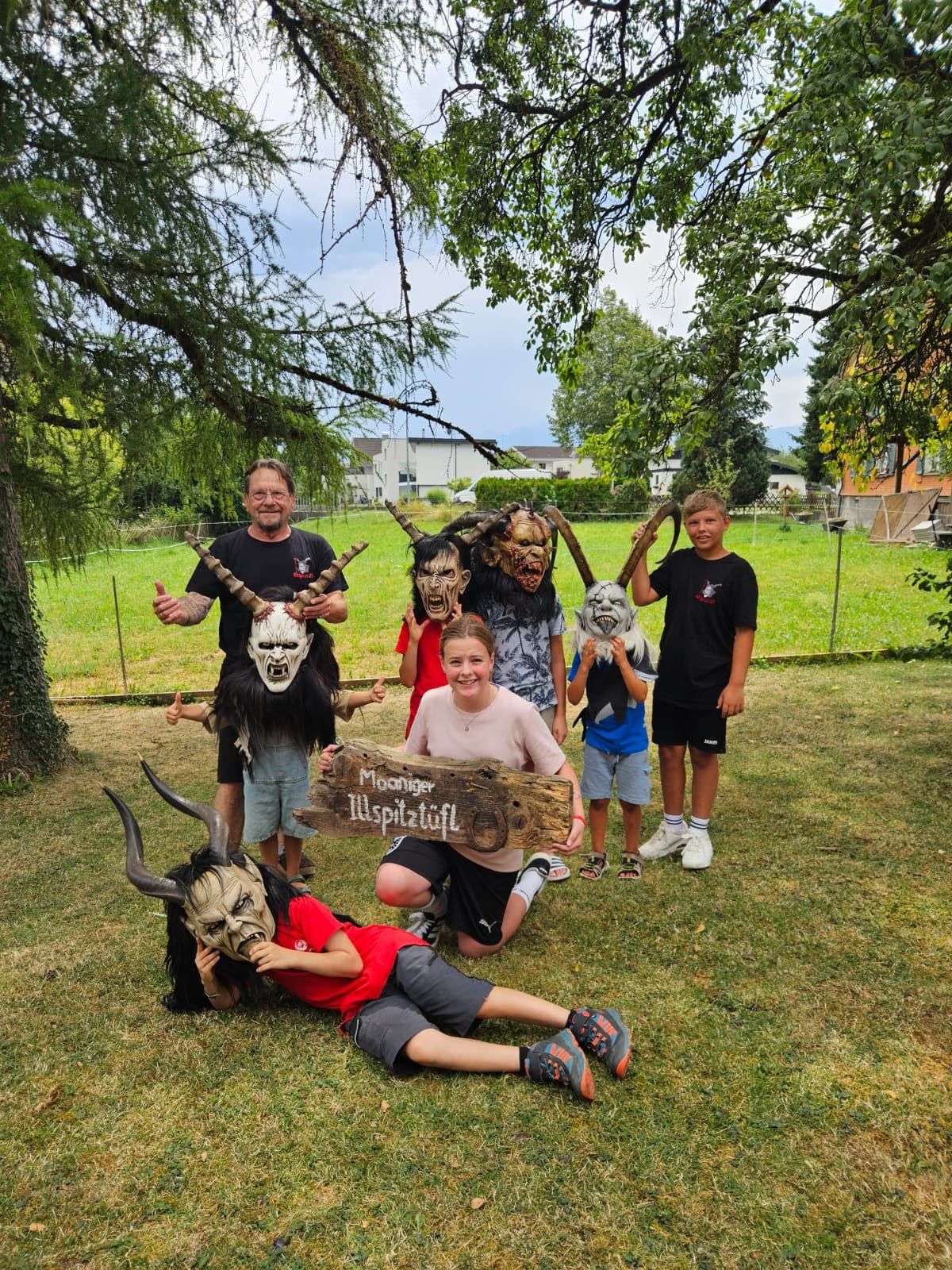 A group of people, including children, are posing for a photo in a grassy field. They are wearing masks with horns and holding a wooden sign that says 'Moosanger'. The background shows a fence and some buildings.